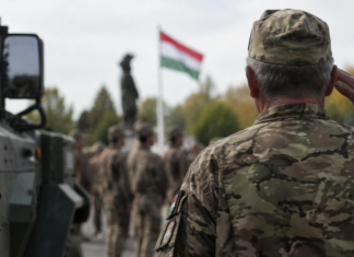 Idősebb magyar katona tiszteleg egy katonai alakulat előtt, háttérben magyar zászlóval és katonai járművel.